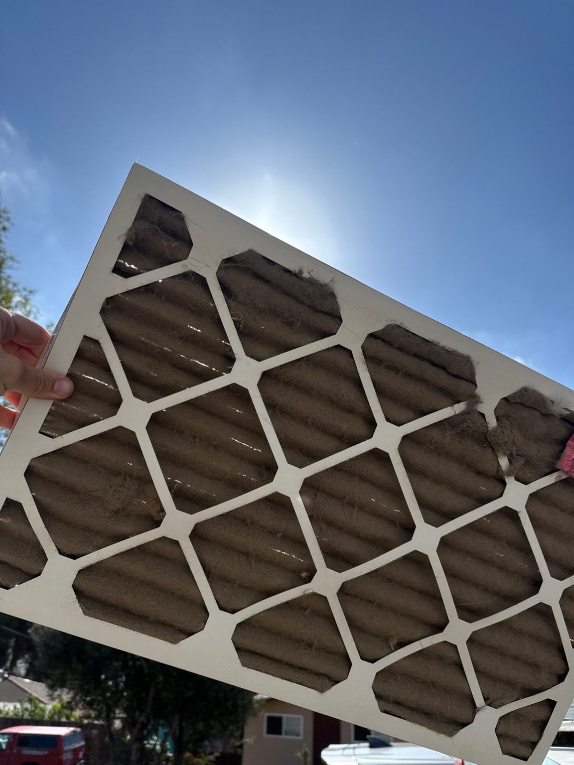 Severely clogged pleated HVAC air filter held against a blue sky, showing how dust buildup blocks airflow to the AC.