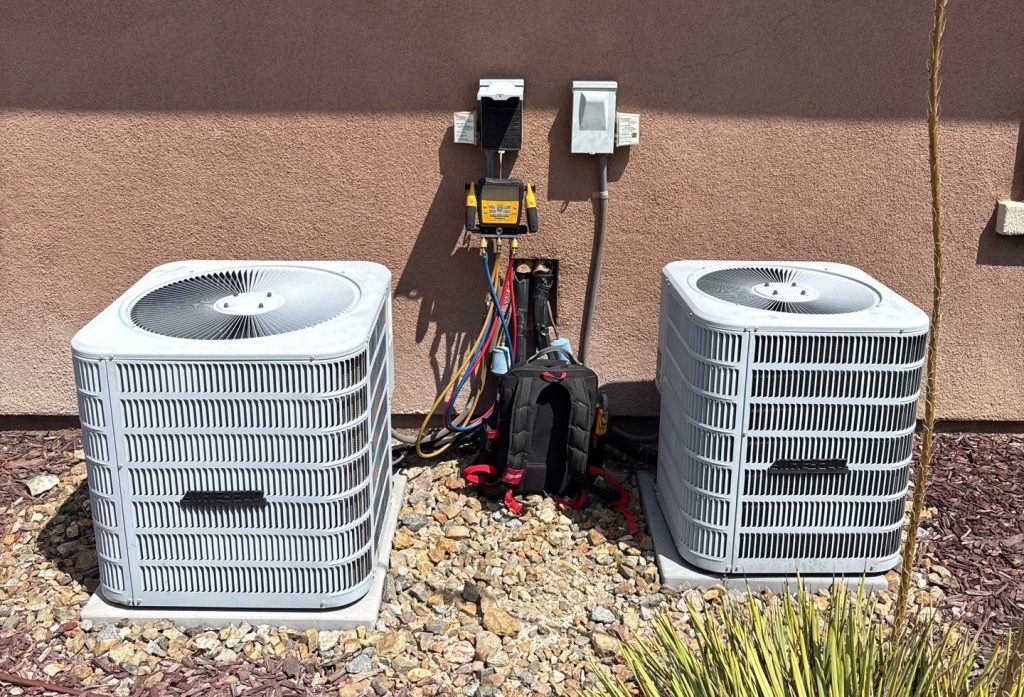 Two residential AC condenser units side by side at a San Diego home with a refrigerant gauge and HVAC technician's tool bag set up between them for diagnostic testing