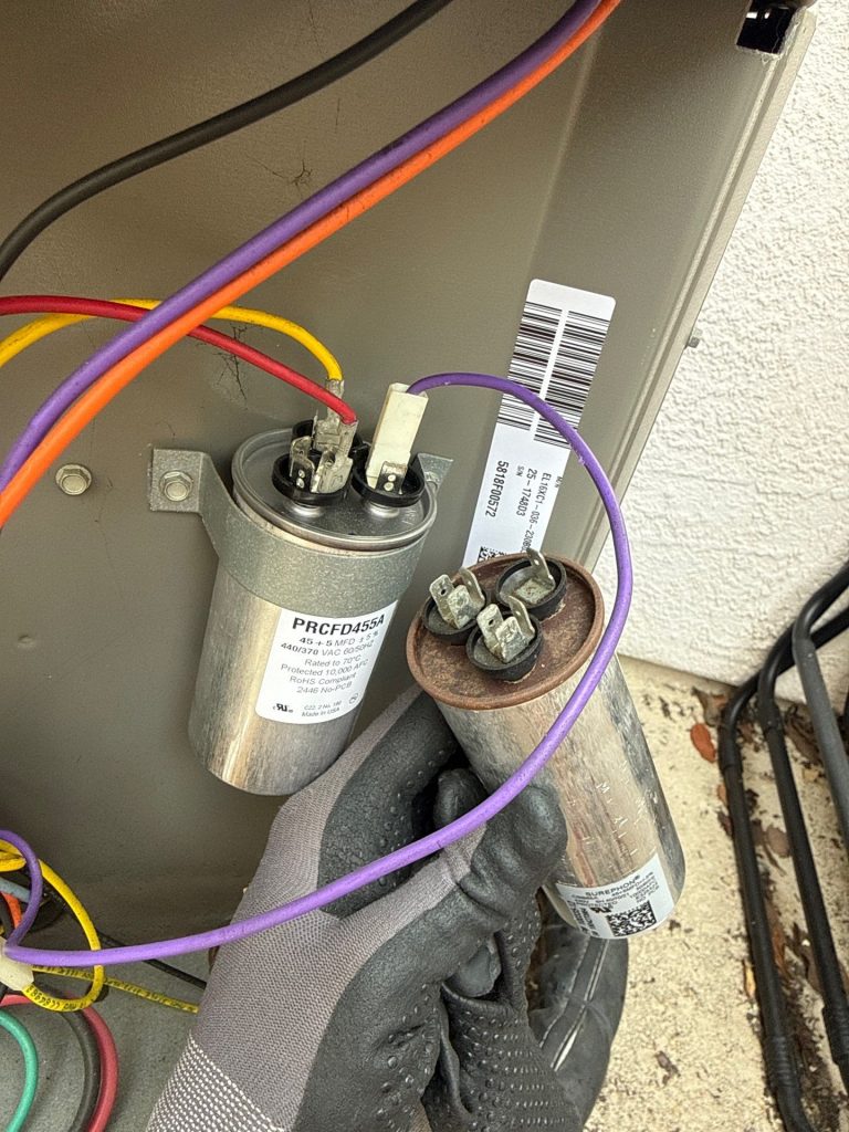 New silver AC capacitor mounted in the condenser next to a rusted, corroded failed capacitor held by a gloved technician.
